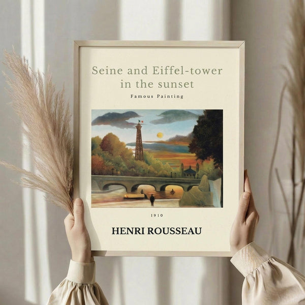 May include: Hands holding framed art print featuring Eiffel Tower and bridge scene in autumn colors with yellow sun, displayed against white curtained window with dried pampas grass. Cream matted print in light wood frame shows naive style painting with green foliage, stone bridge arches, and small figure by water.