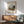 May include: Framed art print featuring Henri Rousseau's painting of the Eiffel Tower and Seine River at sunset, displayed above a mid-century modern wooden credenza with white ceramic lamp. Sage green upholstered armchair with wooden frame sits nearby on light wood floors. Warm natural light creates geometric shadows across white wall in minimalist living room setting.