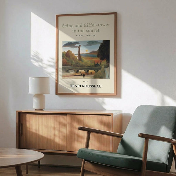 May include: Framed art print featuring Henri Rousseau's painting of the Eiffel Tower and Seine River at sunset, displayed above a mid-century modern wooden credenza with white ceramic lamp. Sage green upholstered armchair with wooden frame sits nearby on light wood floors. Warm natural light creates geometric shadows across white wall in minimalist living room setting.