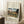 May include: Framed art print leaning against white wall corner featuring Eiffel Tower and Seine river landscape in autumn colors with bridge and trees. Light wood frame on white matting, positioned on textured white rug next to wooden side table with coffee cup. Natural lighting creates soft shadows across the scene.