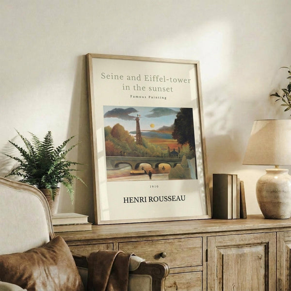 May include: Framed art print featuring Henri Rousseau's painting of the Eiffel Tower and Seine River at sunset, displayed on a rustic wooden sideboard alongside books, a ceramic vase, fern plant, and table lamp in a cozy living room setting with neutral tones and warm lighting.