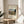 May include: Framed poster displaying a painting of the Eiffel Tower and Seine River at sunset, featuring warm autumn colors with trees, a stone bridge, and small figures. The cream-colored print shows "Seine and Eiffel-tower in the sunset Famous Painting 1910 HENRI ROUSSEAU" text. Displayed on a round wooden side table next to a beige upholstered chair, with plant leaves visible and natural sunlight creating soft shadows on white walls.