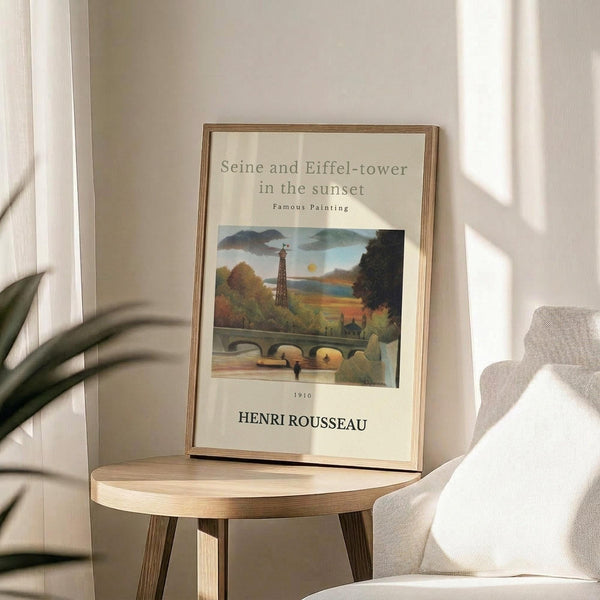 May include: Framed poster displaying a painting of the Eiffel Tower and Seine River at sunset, featuring warm autumn colors with trees, a stone bridge, and small figures. The cream-colored print shows "Seine and Eiffel-tower in the sunset Famous Painting 1910 HENRI ROUSSEAU" text. Displayed on a round wooden side table next to a beige upholstered chair, with plant leaves visible and natural sunlight creating soft shadows on white walls.