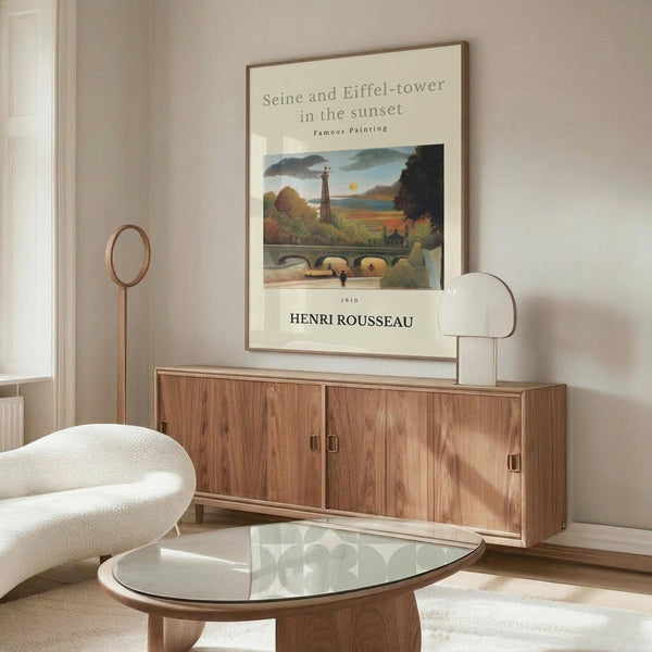 May include: Framed art print featuring Henri Rousseau's 1910 painting of the Seine River and Eiffel Tower at sunset, displayed above a wooden sideboard with sliding doors in a modern living room. White upholstered chair, glass-top oval coffee table, and minimalist decor complete the neutral-toned interior scene.