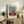 May include: Framed art print displaying Henri Rousseau's painting of a blonde child in a red polka-dotted dress holding a doll, set against a blue sky and green ground. The beige matted print sits on a rustic wooden sideboard alongside ferns, books, ceramic vases, and a table lamp in a neutral-toned living room.
