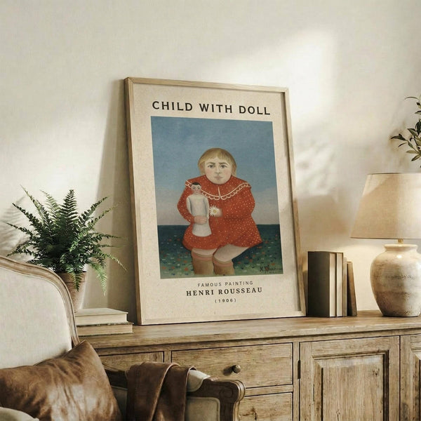 May include: Framed art print displaying Henri Rousseau's painting of a blonde child in a red polka-dotted dress holding a doll, set against a blue sky and green ground. The beige matted print sits on a rustic wooden sideboard alongside ferns, books, ceramic vases, and a table lamp in a neutral-toned living room.