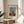 May include: Framed art print displaying a naive-style painting of a blonde child in a red polka-dotted dress holding a white doll, set against a blue background. The poster has cream matting with black text reading "CHILD WITH DOLL" and "FAMOUS PAINTING HENRI ROUSSEAU (1906)". Shows thin gold frame on a round wooden side table next to a beige upholstered chair, with plant leaves visible and natural sunlight casting shadows on white walls.
