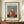 May include: Framed art print of Henri Rousseau's Child with Doll painting displayed on wooden sideboard. Shows young child in red polka dot dress holding white doll against blue background. Cream matting with brown frame. White ceramic vase and stacked books nearby. Neutral wall with white molding visible.