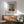 May include: Framed poster print featuring a young child in a red dress holding a white doll, displayed above a wooden sideboard with white ceramic lamp in a modern living room with mint green upholstered chair and natural lighting creating geometric shadows on white wall.