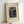 May include: Framed art print leaning against white wall showing Henri Rousseau's 1909 painting "The Muse Inspiring the Poet" featuring two figures in dark clothing standing among lush jungle foliage. Print has cream matting with black text identifying the artist, date, and title. Light wooden frame casts soft shadow on wall. Partial view of hand in corner for scale reference.