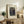 May include: Framed art print of Henri Rousseau's 1909 painting showing two figures in dark clothing surrounded by lush jungle foliage, displayed on a rustic wooden sideboard alongside a fern plant, stacked books, and ceramic pottery in a warm, neutral-toned living room setting.