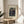 May include: Framed art print featuring Henri Rousseau's 1909 painting "The Muse Inspiring the Poet" displayed on a round wooden side table next to a cream upholstered chair. The vintage-style poster shows three figures in a lush jungle setting with dense foliage. Natural sunlight streams across the neutral-toned room, casting soft shadows from a plant visible in the corner.