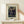 May include: Framed art print displaying Henri Rousseau's 1909 painting featuring three figures in dark clothing standing among lush green tropical foliage. The vintage-style poster has cream matting and thin wooden frame, positioned on a light wood shelf against white wall. A hand reaches toward the frame from the right side, with a small white wrapped gift box sitting on the shelf below the artwork.