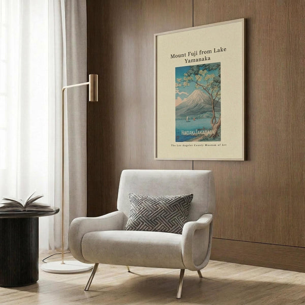 May include: Vintage-style poster of Mount Fuji with cherry blossom tree and lake in soft blues and pastels, displayed in light wooden frame on wood-paneled wall above beige upholstered armchair with geometric patterned pillow, alongside brass floor lamp and dark side table with open book