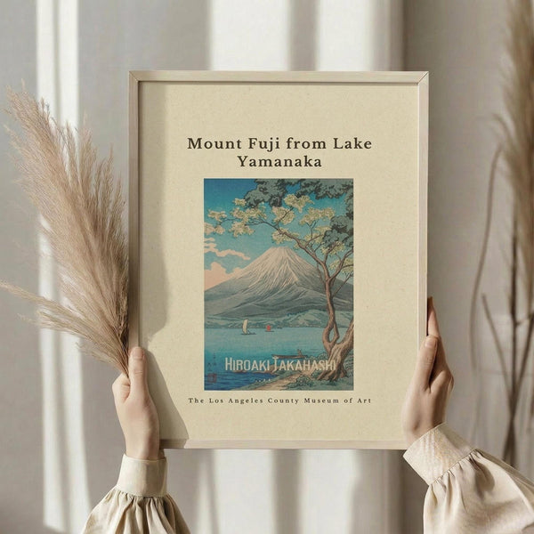 May include: Person in cream sweater holding framed vintage Japanese woodblock print showing Mount Fuji viewed across Lake Yamanaka, with cherry blossom tree in foreground and small boats on blue water. Print features soft blues, greens and pink tones in traditional ukiyo-e style. Light wooden frame against cream matting. Dried pampas grass stems visible to left, neutral curtained background.
