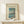 May include: Framed vintage-style Japanese woodblock print showing Mount Fuji across a blue lake with a twisted tree in foreground and small boats on water. Print has cream matting in light wood frame, displayed on white wooden console table next to small white gift box with twine and greenery sprig against off-white wall with soft natural lighting.