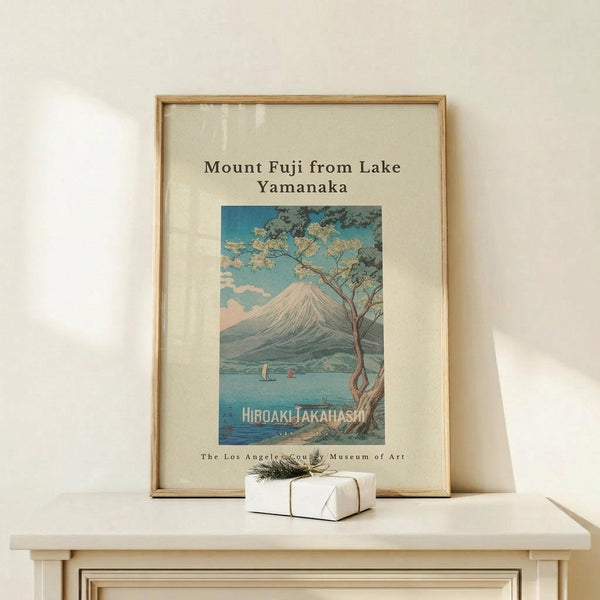 May include: Framed vintage-style Japanese woodblock print showing Mount Fuji across a blue lake with a twisted tree in foreground and small boats on water. Print has cream matting in light wood frame, displayed on white wooden console table next to small white gift box with twine and greenery sprig against off-white wall with soft natural lighting.