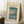 May include: Framed vintage-style Japanese woodblock print poster showing Mount Fuji viewed across Lake Yamanaka, with a twisted tree in foreground and small boats on blue water. The artwork features soft blue skies, snow-capped mountain peak, and traditional artistic styling. Displayed in light wood frame leaning against white wall corner, next to coffee cup on wooden block and cream textured throw blanket.