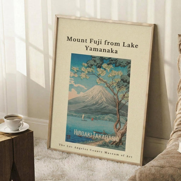 May include: Framed vintage-style Japanese woodblock print poster showing Mount Fuji viewed across Lake Yamanaka, with a twisted tree in foreground and small boats on blue water. The artwork features soft blue skies, snow-capped mountain peak, and traditional artistic styling. Displayed in light wood frame leaning against white wall corner, next to coffee cup on wooden block and cream textured throw blanket.