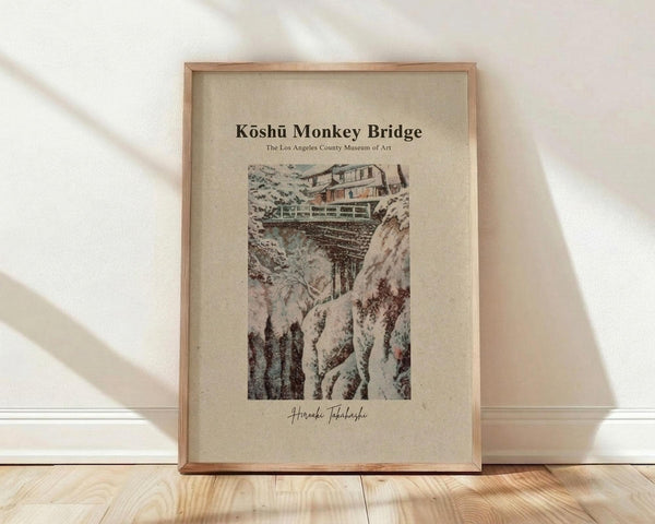 May include: Framed vintage Japanese woodblock print showing a snow-covered bridge spanning a rocky gorge with traditional buildings above. The artwork features muted earth tones and delicate linework typical of ukiyo-e style. Displayed in a light wooden frame against white fabric backdrop with natural shadows on pale wooden flooring.