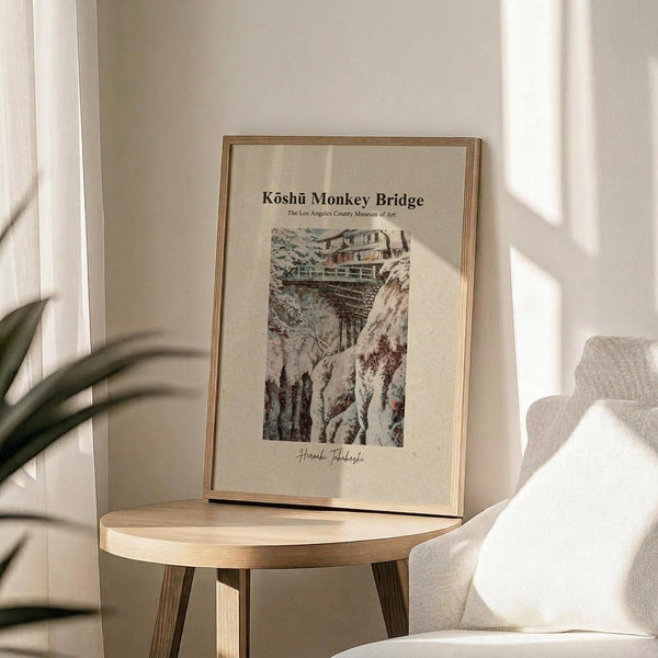 May include: Framed vintage Japanese woodblock print depicting a snow-covered mountain bridge scene displayed on a light wood circular side table. The artwork shows rocky cliffs, traditional architecture, and small figures crossing a bridge in muted earth tones. Soft natural lighting creates shadows across cream-colored walls, with a beige upholstered chair and green plant leaves visible in the minimalist room setting.