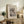 May include: Framed vintage-style art print featuring a traditional Japanese bridge over a rocky gorge with buildings, displayed on rustic wooden console table alongside fern plant, ceramic vase, and stacked books in neutral-toned living room setting with cream upholstered chair and warm lighting