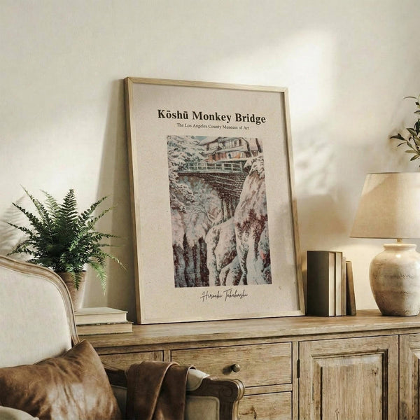 May include: Framed vintage-style art print featuring a traditional Japanese bridge over a rocky gorge with buildings, displayed on rustic wooden console table alongside fern plant, ceramic vase, and stacked books in neutral-toned living room setting with cream upholstered chair and warm lighting