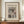 May include: Framed art print displayed on wooden sideboard showing Japanese woodblock-style winter landscape with snow-covered bridge and rocky cliffs in muted earth tones. Brown frame sits beside small white ceramic vase and stacked books against cream paneled wall. Print features museum attribution text and artist signature at bottom.