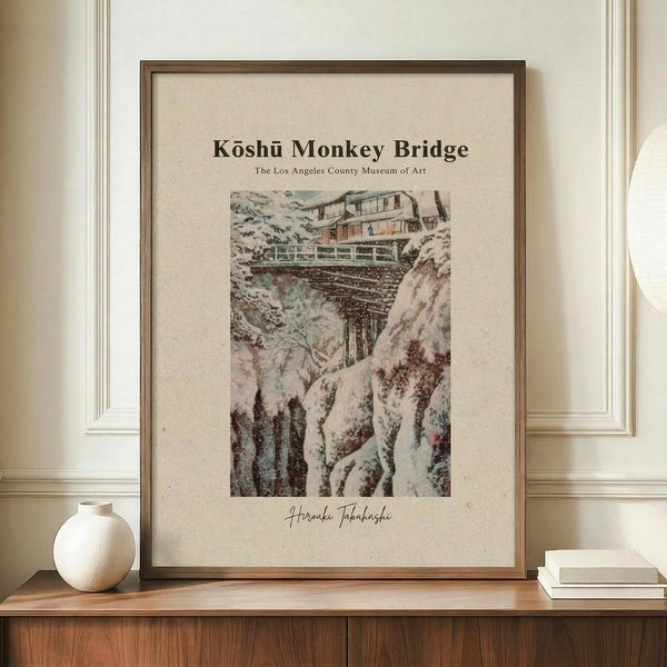 May include: Framed art print displayed on wooden sideboard showing Japanese woodblock-style winter landscape with snow-covered bridge and rocky cliffs in muted earth tones. Brown frame sits beside small white ceramic vase and stacked books against cream paneled wall. Print features museum attribution text and artist signature at bottom.