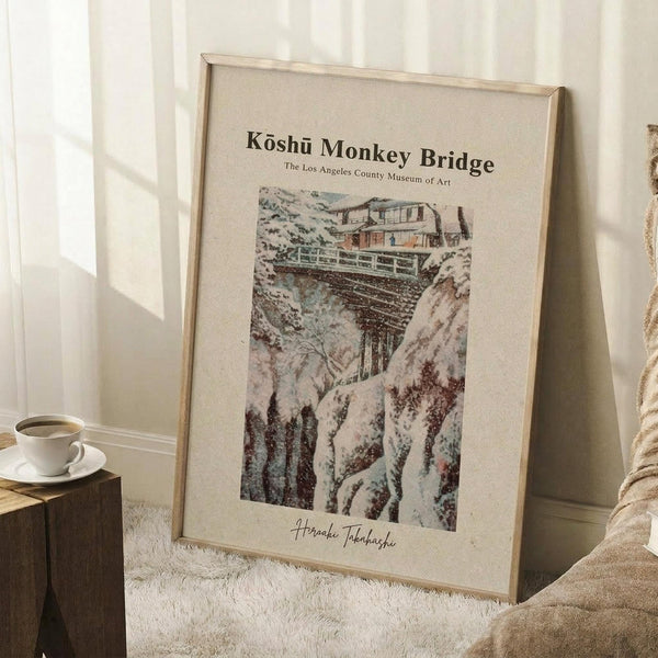 May include: A framed vintage-style Japanese woodblock print showing a snow-covered mountain bridge scene with traditional buildings, displayed in a light wood frame leaning against white wainscoting. A white coffee cup sits on a wooden block nearby on a cream textured rug, with soft natural lighting creating shadows on the wall.