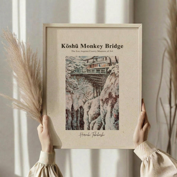 May include: A framed vintage-style Japanese woodblock print showing a wooden bridge spanning a rocky gorge with traditional buildings above. The artwork features muted earth tones and depicts a winter or snowy mountain landscape. Held by hands in cream-colored clothing with gathered sleeves, alongside dried pampas grass stems. Set against soft neutral curtains in natural window lighting. The print has aged, cream-colored matting in a simple wooden frame.
