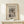 May include: Framed vintage-style Japanese woodblock print showing a snow-covered bridge spanning a rocky gorge, with traditional buildings visible above. The artwork features muted earth tones and depicts a winter mountain landscape. Displayed on a light wooden shelf against a cream wall, with a small wrapped gift box with twine and evergreen sprig positioned in front.