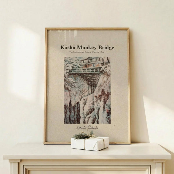 May include: Framed vintage-style Japanese woodblock print showing a snow-covered bridge spanning a rocky gorge, with traditional buildings visible above. The artwork features muted earth tones and depicts a winter mountain landscape. Displayed on a light wooden shelf against a cream wall, with a small wrapped gift box with twine and evergreen sprig positioned in front.
