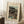 May include: Framed vintage Japanese woodblock print showing two figures in traditional dress walking beneath a large tree near water at dusk or dawn. The artwork features muted earth tones with teal blue water and depicts a serene landscape scene. The cream matted print is displayed in a thin wooden frame, leaning against white paneled walls on a textured white rug. A wooden side table with white coffee cup and saucer sits nearby, with soft beige fabric visible at the edge of the scene.