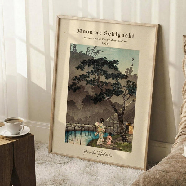 May include: Framed vintage Japanese woodblock print showing two figures in traditional dress walking beneath a large tree near water at dusk or dawn. The artwork features muted earth tones with teal blue water and depicts a serene landscape scene. The cream matted print is displayed in a thin wooden frame, leaning against white paneled walls on a textured white rug. A wooden side table with white coffee cup and saucer sits nearby, with soft beige fabric visible at the edge of the scene.