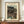 May include: Framed Japanese woodblock print poster displaying a moonlit evening scene with figures in traditional dress walking near water and a large tree, featuring muted green and brown tones. The vintage-style artwork is mounted in a brown wooden frame, displayed on a walnut credenza beside a white ceramic vase and stacked books against cream wainscoted walls.