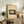 May include: Framed vintage Japanese woodblock print showing moonlit rural landscape with trees, houses and mountains in sepia tones, displayed on rustic wooden sideboard alongside fern plant, ceramic vase, and stacked books in neutral-toned living room setting