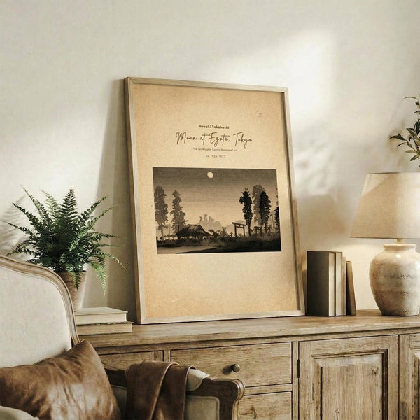 May include: Framed vintage Japanese woodblock print showing moonlit rural landscape with trees, houses and mountains in sepia tones, displayed on rustic wooden sideboard alongside fern plant, ceramic vase, and stacked books in neutral-toned living room setting