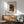 May include: Japanese woodblock print poster showing moonlit evening scene with figures by water and traditional bridge, displayed in light wood frame above mid-century credenza. Sage green upholstered armchair with wooden arms sits nearby, white ceramic table lamp on credenza, warm sunlight casting geometric shadows across white wall.
