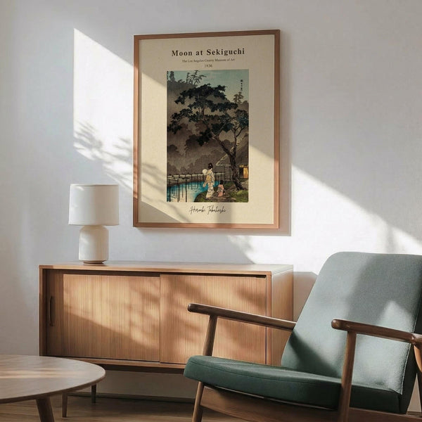 May include: Japanese woodblock print poster showing moonlit evening scene with figures by water and traditional bridge, displayed in light wood frame above mid-century credenza. Sage green upholstered armchair with wooden arms sits nearby, white ceramic table lamp on credenza, warm sunlight casting geometric shadows across white wall.