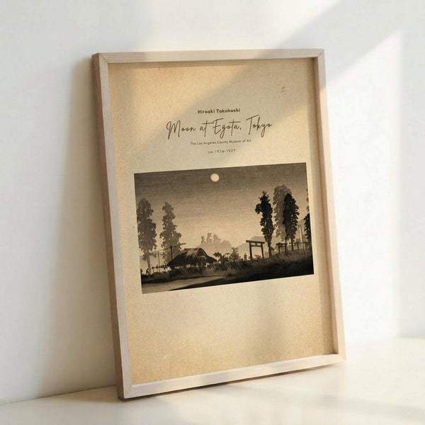 May include: Framed vintage Japanese woodblock print reproduction showing a moonlit evening landscape with silhouetted trees, traditional buildings, and torii gate against a dusky sky. The cream-colored artwork is displayed in a light wooden frame, leaning against a white wall corner with soft natural lighting creating gentle shadows.