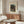May include: Japanese woodblock print poster in thin frame hanging above wooden mid-century credenza in minimalist living room. Print shows moonlit evening scene with figures under tree near water and bridge in muted blues and greens. White upholstered chair, glass coffee table, and white table lamp complete the neutral-toned interior design mockup.