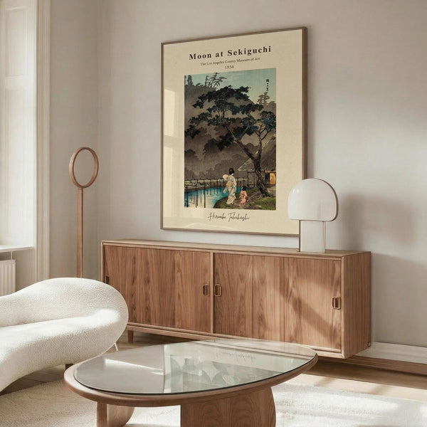 May include: Japanese woodblock print poster in thin frame hanging above wooden mid-century credenza in minimalist living room. Print shows moonlit evening scene with figures under tree near water and bridge in muted blues and greens. White upholstered chair, glass coffee table, and white table lamp complete the neutral-toned interior design mockup.