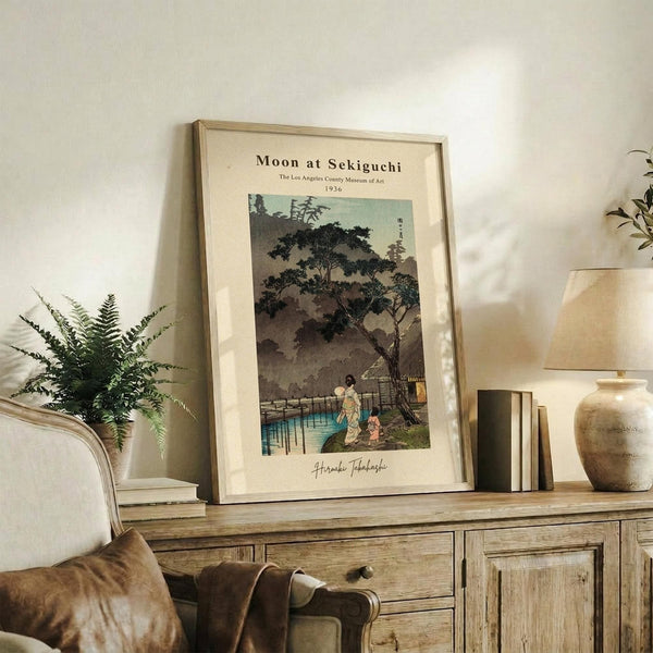 May include: Framed vintage Japanese woodblock print showing figures under moonlit trees by water, displayed on rustic wooden sideboard beside fern plant, lamp, ceramic vase and books in neutral-toned living room setting