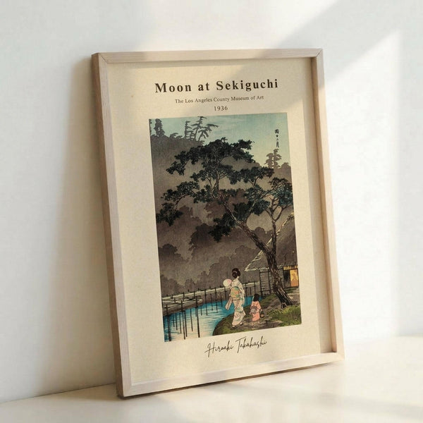 May include: Framed vintage Japanese woodblock print showing a moonlit evening scene at Sekiguchi with figures in traditional dress walking near water, surrounded by large trees and traditional architecture. The artwork features muted earth tones with touches of blue and green, displayed in a light wood frame against a white wall and surface.