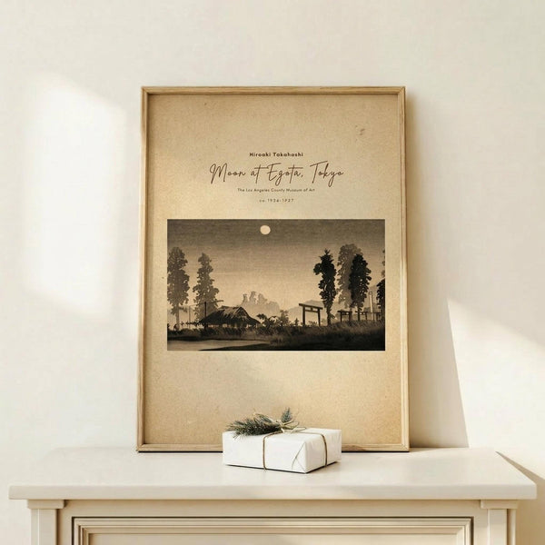 May include: Framed vintage-style Japanese woodblock print showing moonlit rural landscape with silhouetted trees, traditional buildings, and torii gate under full moon. Sepia-toned artwork in light wooden frame displayed on cream wooden shelf beside small white gift box tied with twine and evergreen sprig against neutral wall.