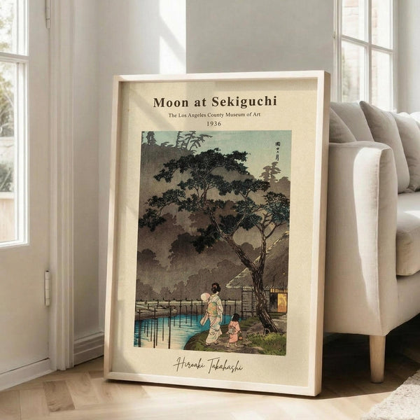 May include: Framed vintage Japanese woodblock print showing moonlit evening scene with figures walking near water and traditional buildings under large tree. Print features muted greens, blues, and earth tones with museum exhibition text at top. Light wood frame leans against white wall next to beige upholstered chair in bright, modern room with hardwood floors and large windows.