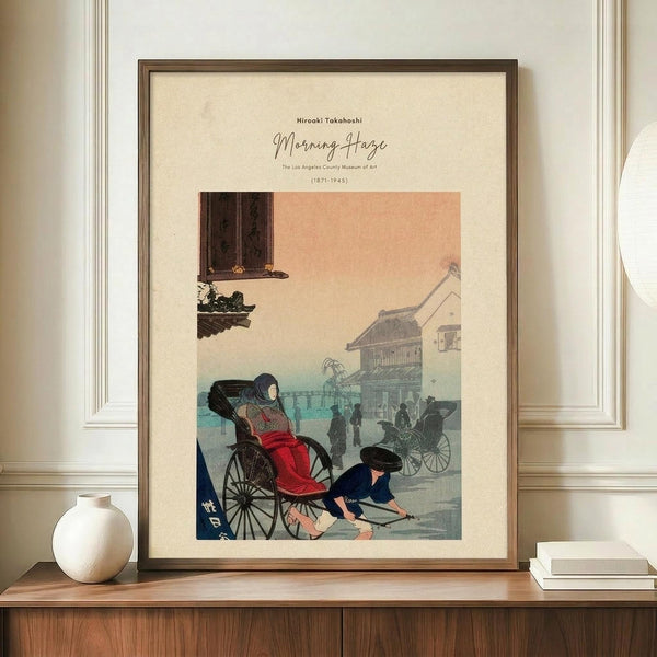 May include: Framed vintage Japanese woodblock print displayed on wooden sideboard beside white ceramic vase and stacked books. The artwork shows a woman in red kimono seated in a rickshaw with a man pulling it through a misty street scene with traditional buildings. Soft peachy-pink sky transitions to blue-green fog, creating atmospheric morning haze effect. Warm beige matting in dark wood frame against cream painted walls with white trim molding.