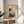 May include: Framed vintage Japanese woodblock print displayed on light wood round side table next to beige upholstered chair. Artwork shows rickshaw pullers in traditional dress on misty street with buildings in background, rendered in muted blues, peach, and earth tones. Natural lighting creates soft shadows across minimalist room setting with plant leaves visible in corner.
