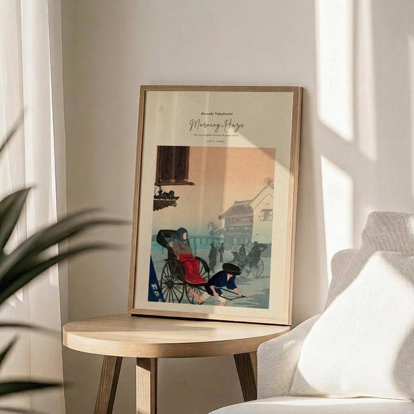 May include: Framed vintage Japanese woodblock print displayed on light wood round side table next to beige upholstered chair. Artwork shows rickshaw pullers in traditional dress on misty street with buildings in background, rendered in muted blues, peach, and earth tones. Natural lighting creates soft shadows across minimalist room setting with plant leaves visible in corner.